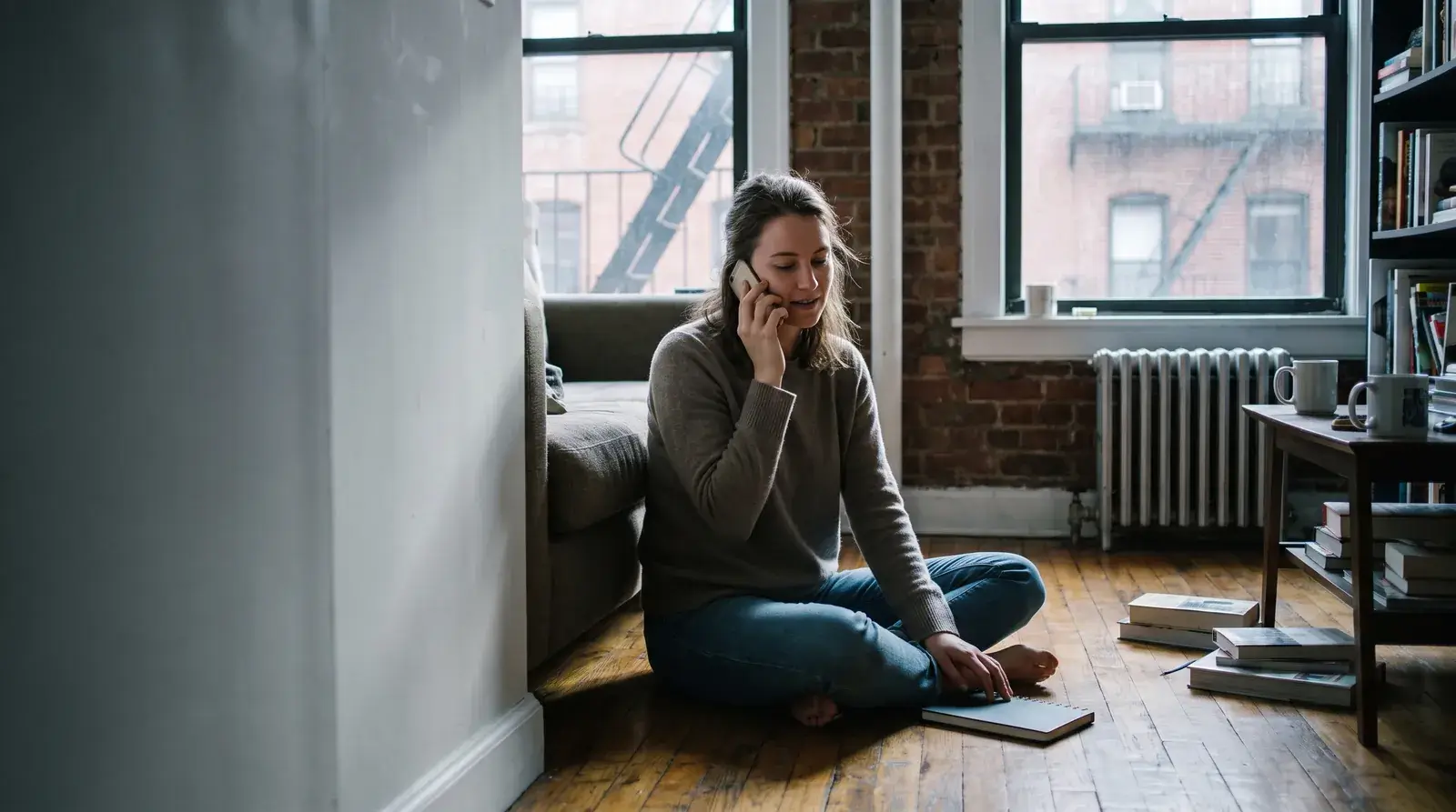 Woman sitting on her apartment floor against the couch, on a phone call in afternoon light