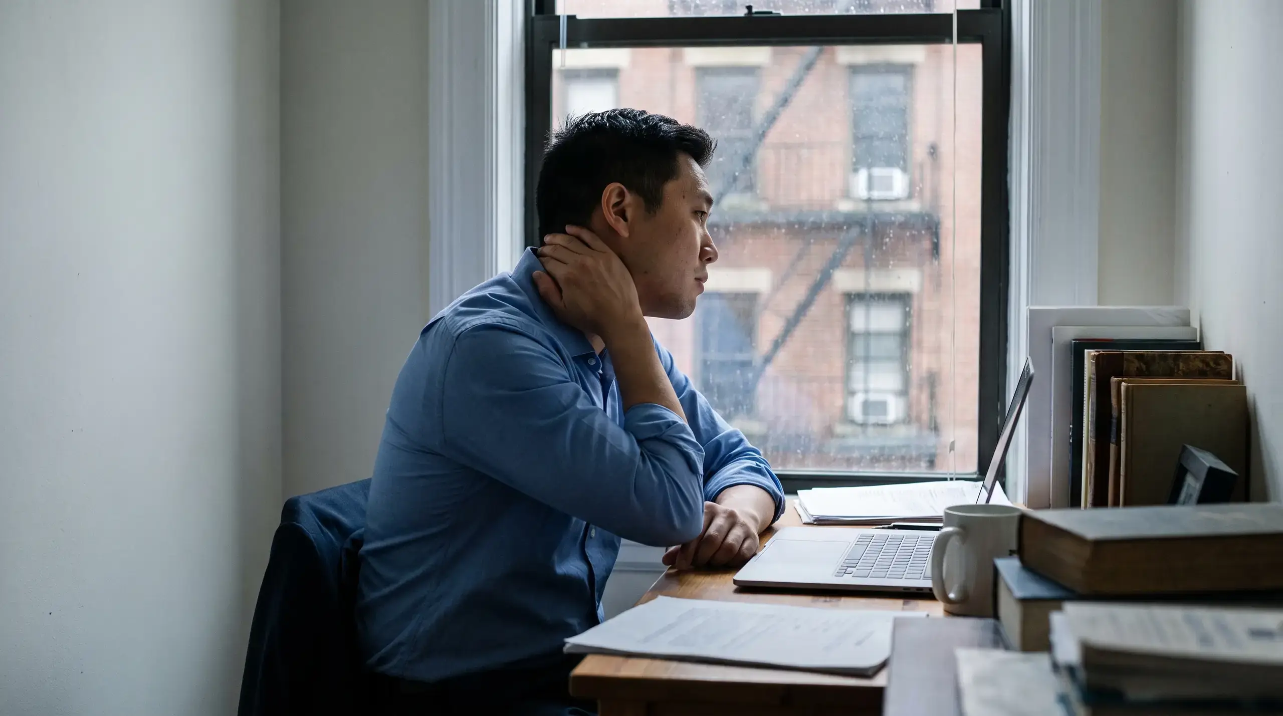 Man in a blue work shirt at a small apartment desk, hand at his neck, looking out a rainy window