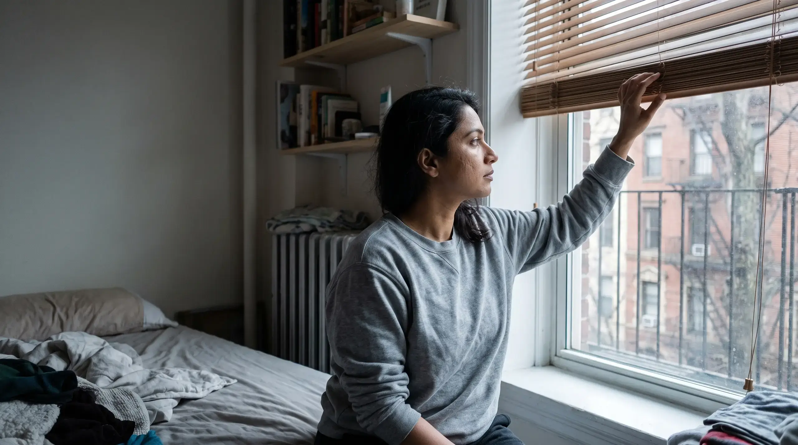 Woman in a gray sweatshirt opening the blinds of her New York bedroom on a slow morning