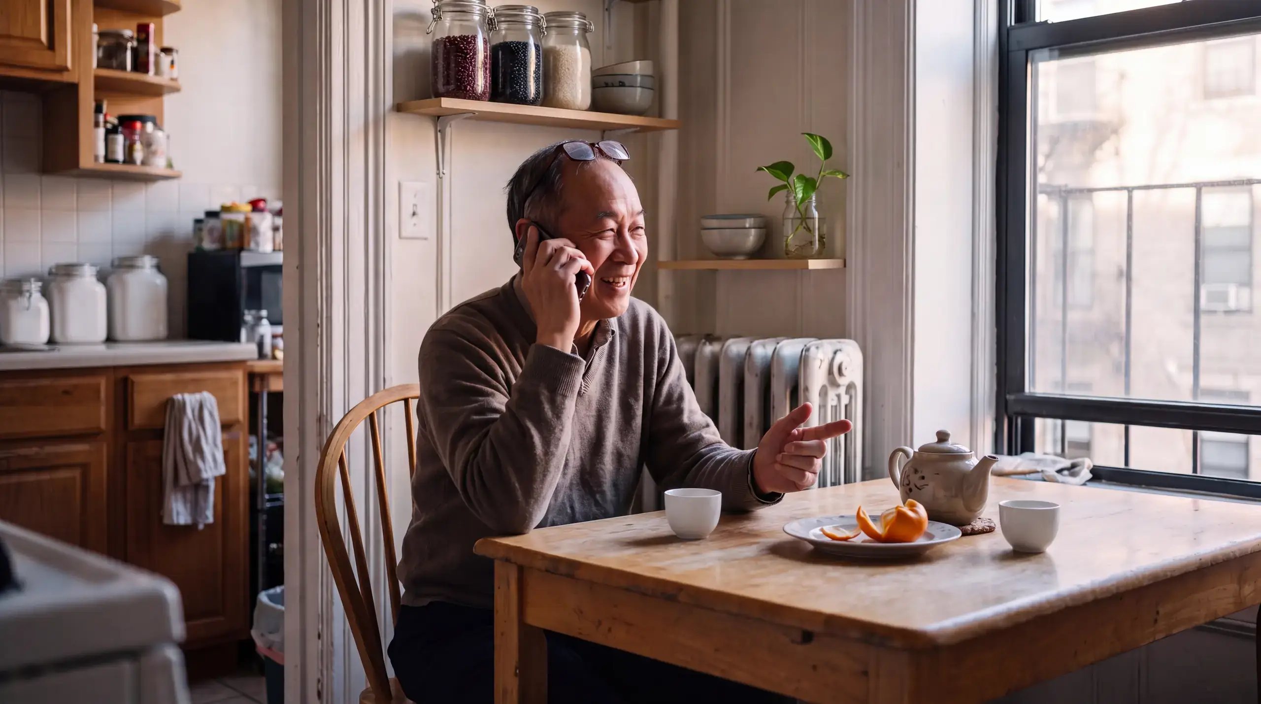 Older man laughing during a phone call at his kitchen table in a New York apartment, tea and an orange beside him